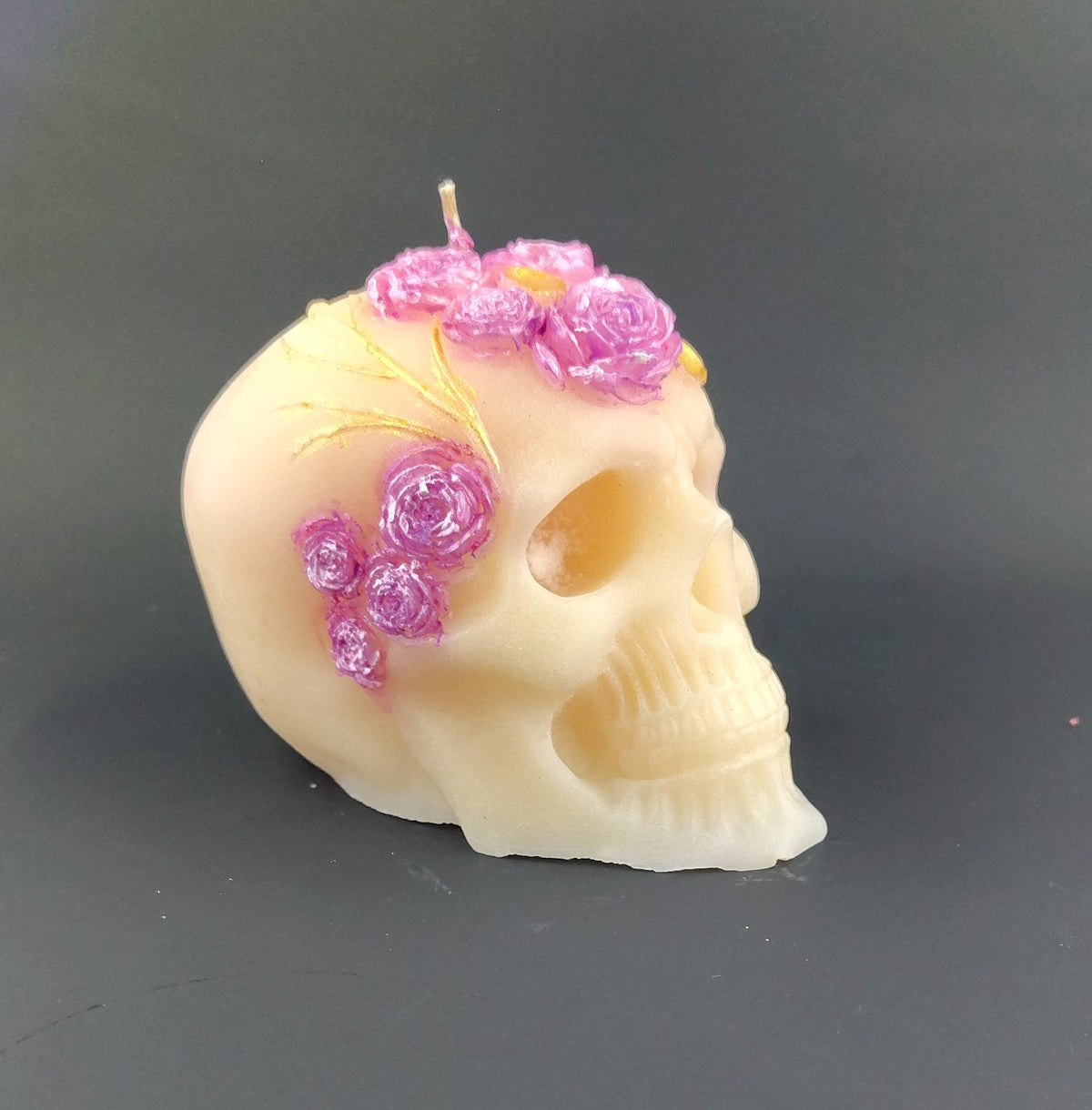 A small skull-shaped candle in an ivory or cream tone, decorated with pink and purple flower details on top, resting on a dark gray surface.