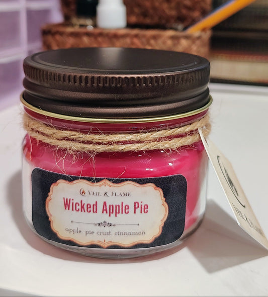 A small glass jar candle with bright red wax, a dark brown metal lid, and a twine-wrapped neck, labeled 'Wicked Apple Pie' by Veil & Flame. The candle is photographed on a white surface with a blurred desk background.