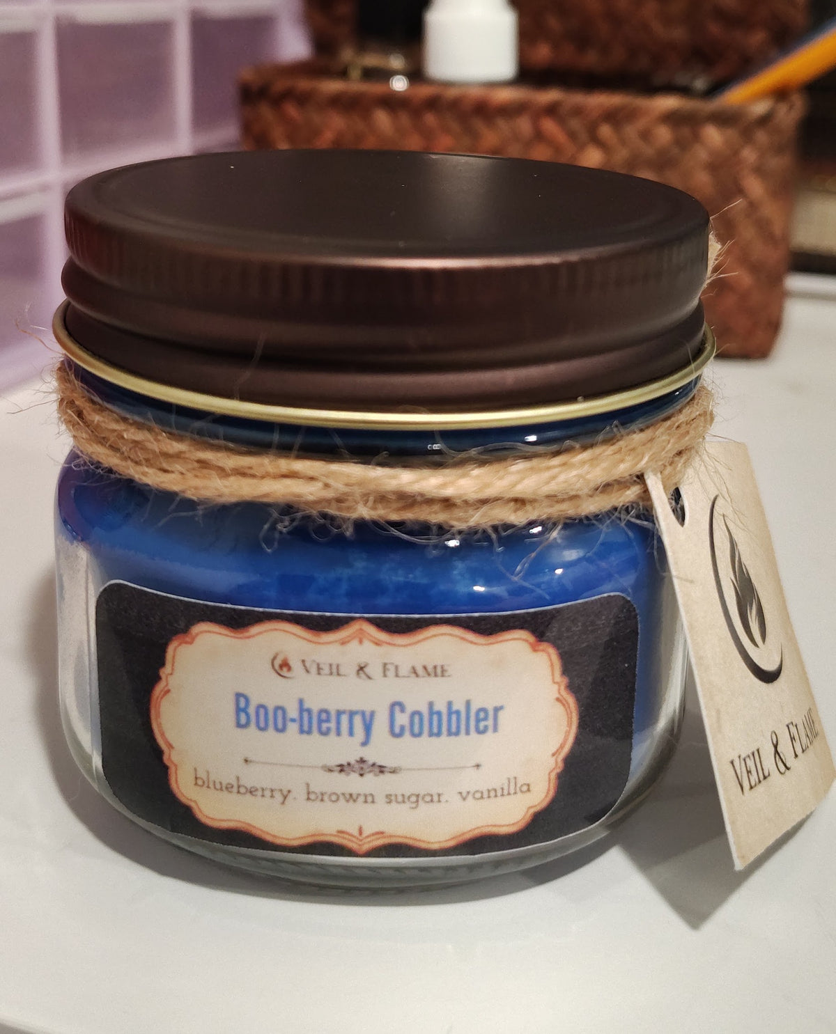 A blue wax candle in a clear glass jar with a dark brown metal lid, wrapped with twine and a hanging tag. The front label reads 'VEIL & FLAME Boo-berry Cobbler'.