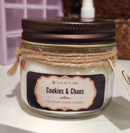 A jar candle labeled 'Cookies & Chaos' by Veil & Flame, with white wax inside a clear glass jar, a dark brown metal lid, and a rustic twine band around the neck.
