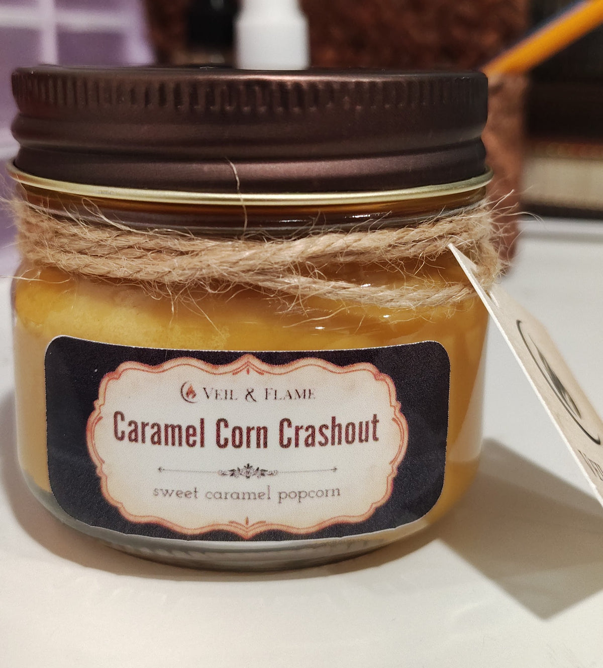 orange colored wax inside a clear glass jar with antique rustic lid. candle is entitled Caramel Corn Crashout and is made by Veil & Flame. Its scent notes are "sweet caramel popcorn". the candle sits on a white desk surface with desk items blurred in the background. 