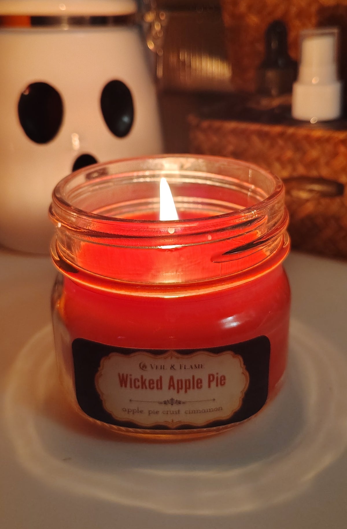 A lit red wax candle in a clear glass jar with a black label reading 'Wicked Apple Pie' by Veil & Flame, set on a white desk with a blurred desk background.