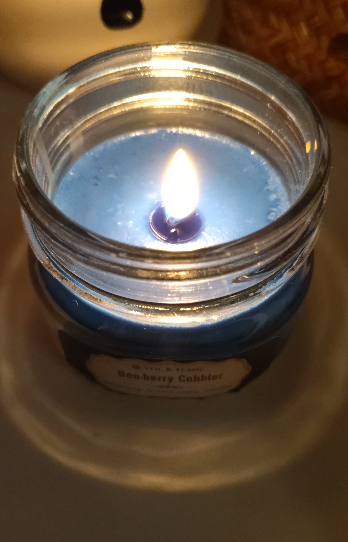 A lit blue candle in a clear glass jar with a visible flame, photographed on a tabletop with a blurred white ceramic mug in the background.