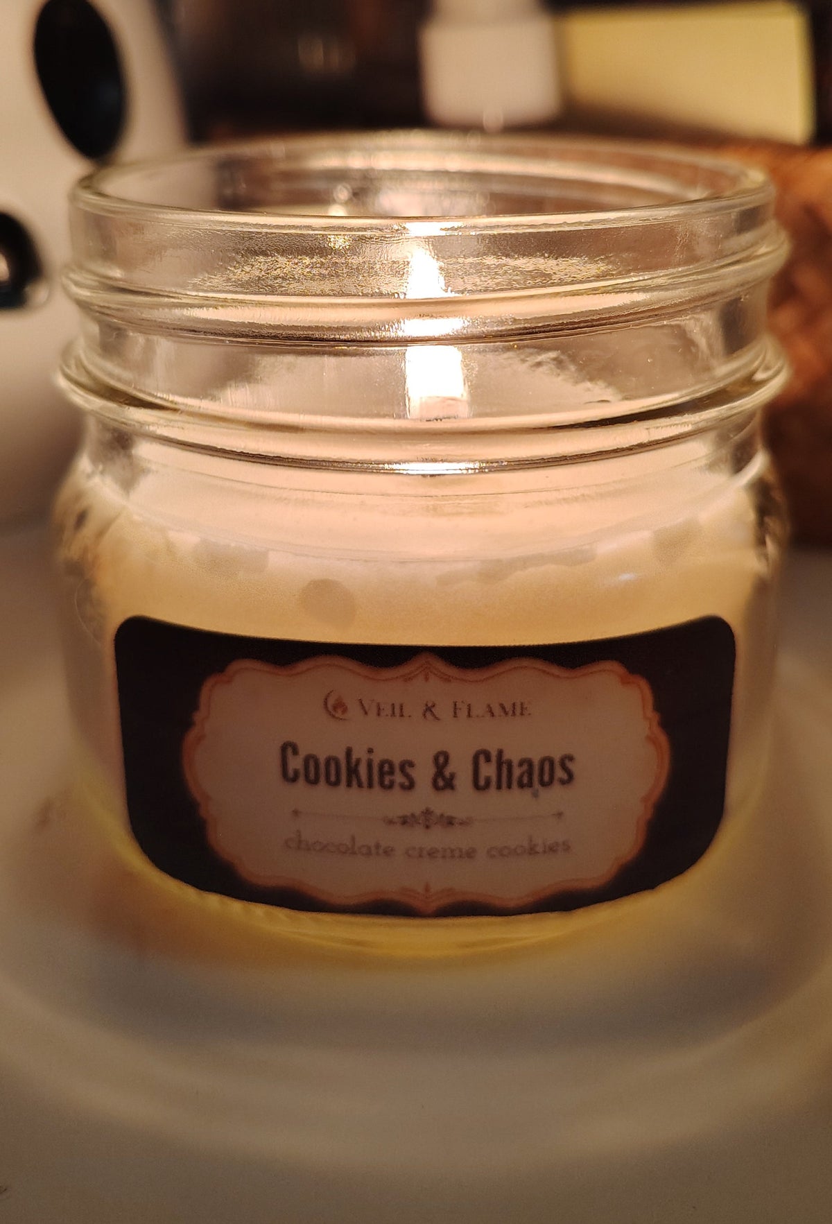 Lit candle in clear glass jar with the name Cookies and Chaos. Logo reads Veil & Flame. Scent notes are chocolate cream cookie. Candle is set on a white surface with a ceramic ghost in the background and a woven basket with some desk items. 