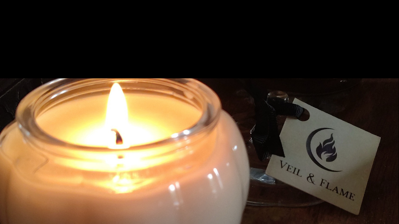 A lit white candle in a clear glass jar with a small hanging tag that reads 'VEIL & FLAME' on a dark wooden surface.