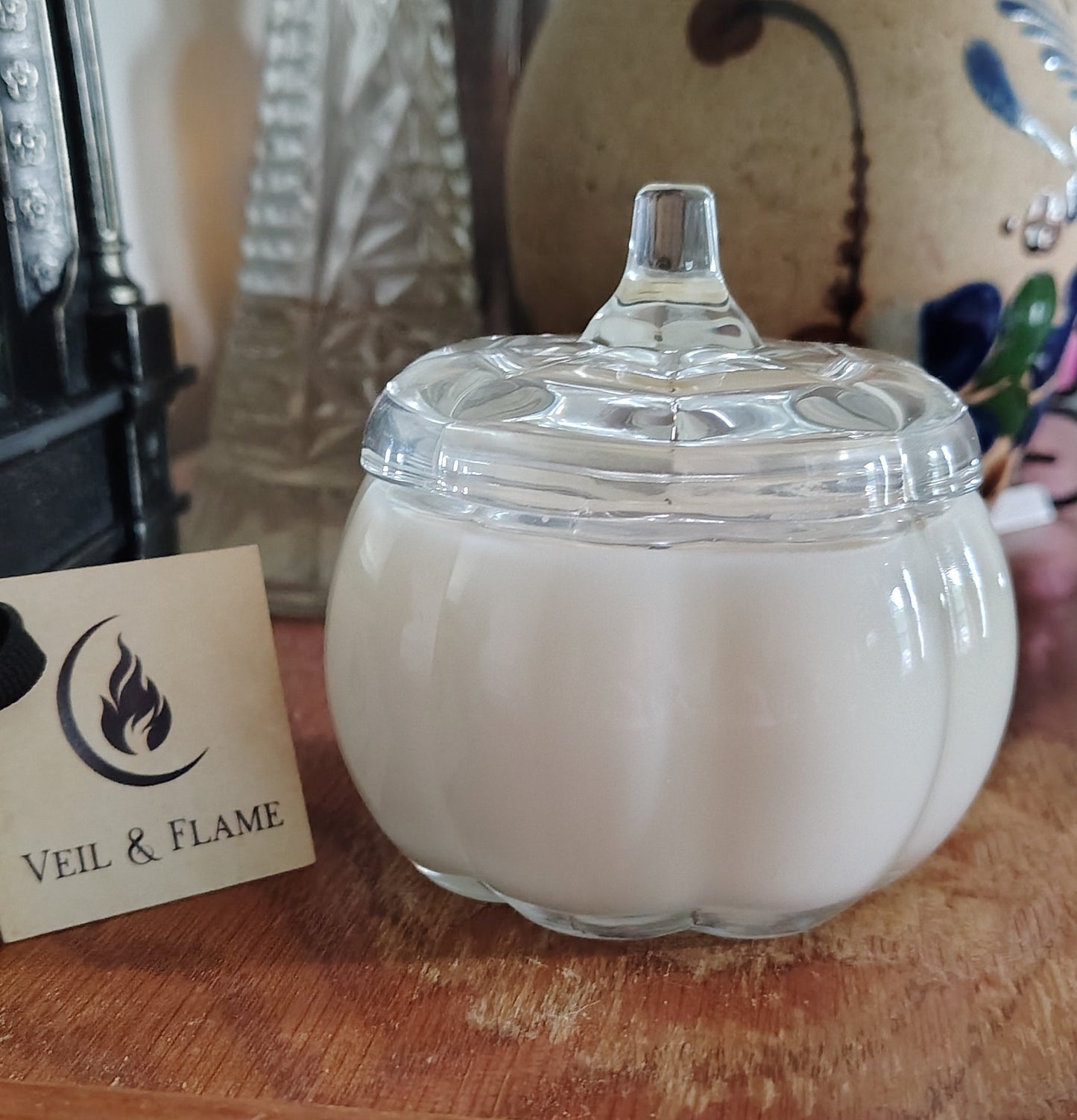 A white wax candle in a clear glass pumpkin-shaped jar with a lid, set on a wooden surface with a branded card in front and decorative vases in the background.