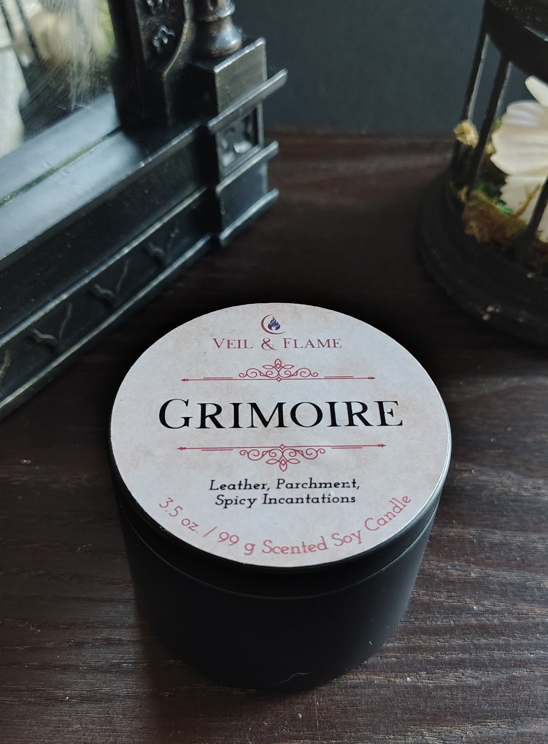 A black round tin of Veil & Flame Grimoire Leather, Parchment, and Spicy Incense Salts candle sits on a dark wooden table next to a vintage mirror.