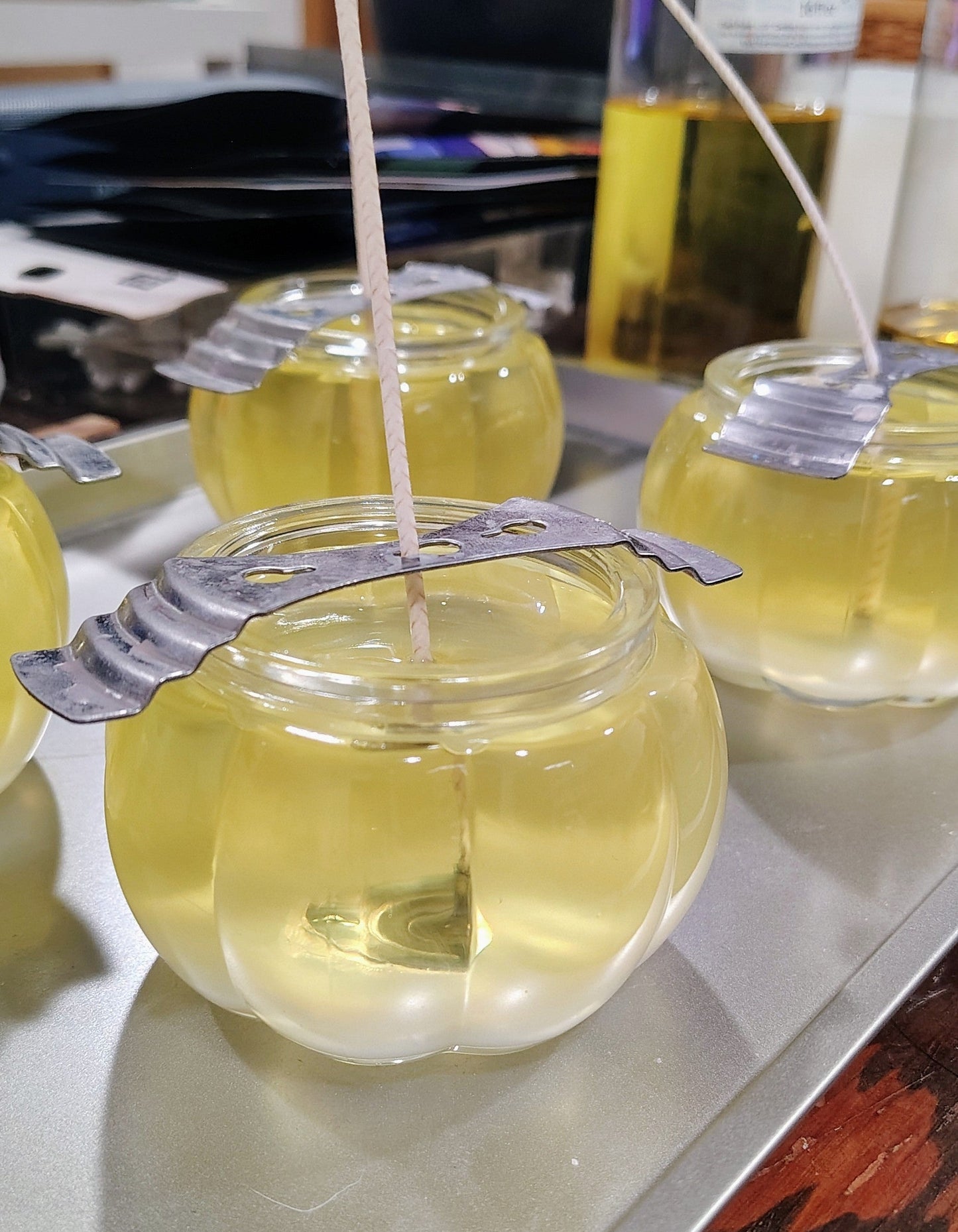 A close-up of a small clear glass jar candle filled with pale yellow wax, sitting on a metal tray. The candle has a metal lid or cover with a wick protruding through it, and other similar candles are partially visible in the background.