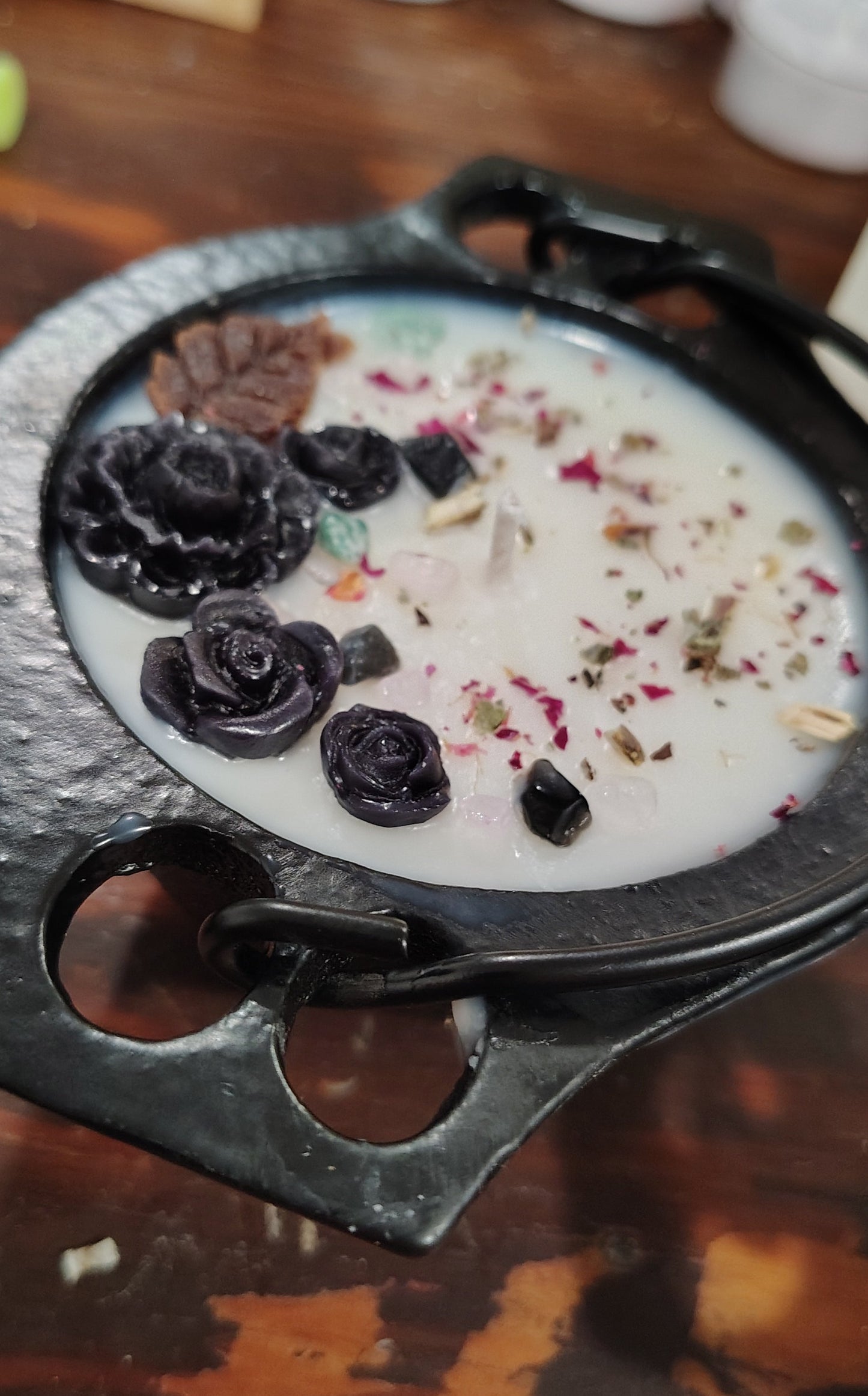 A black cast iron cauldron with decorative handles, filled with a white wax candle topped with dried botanicals and dark rose petals, set on a rustic wooden surface.