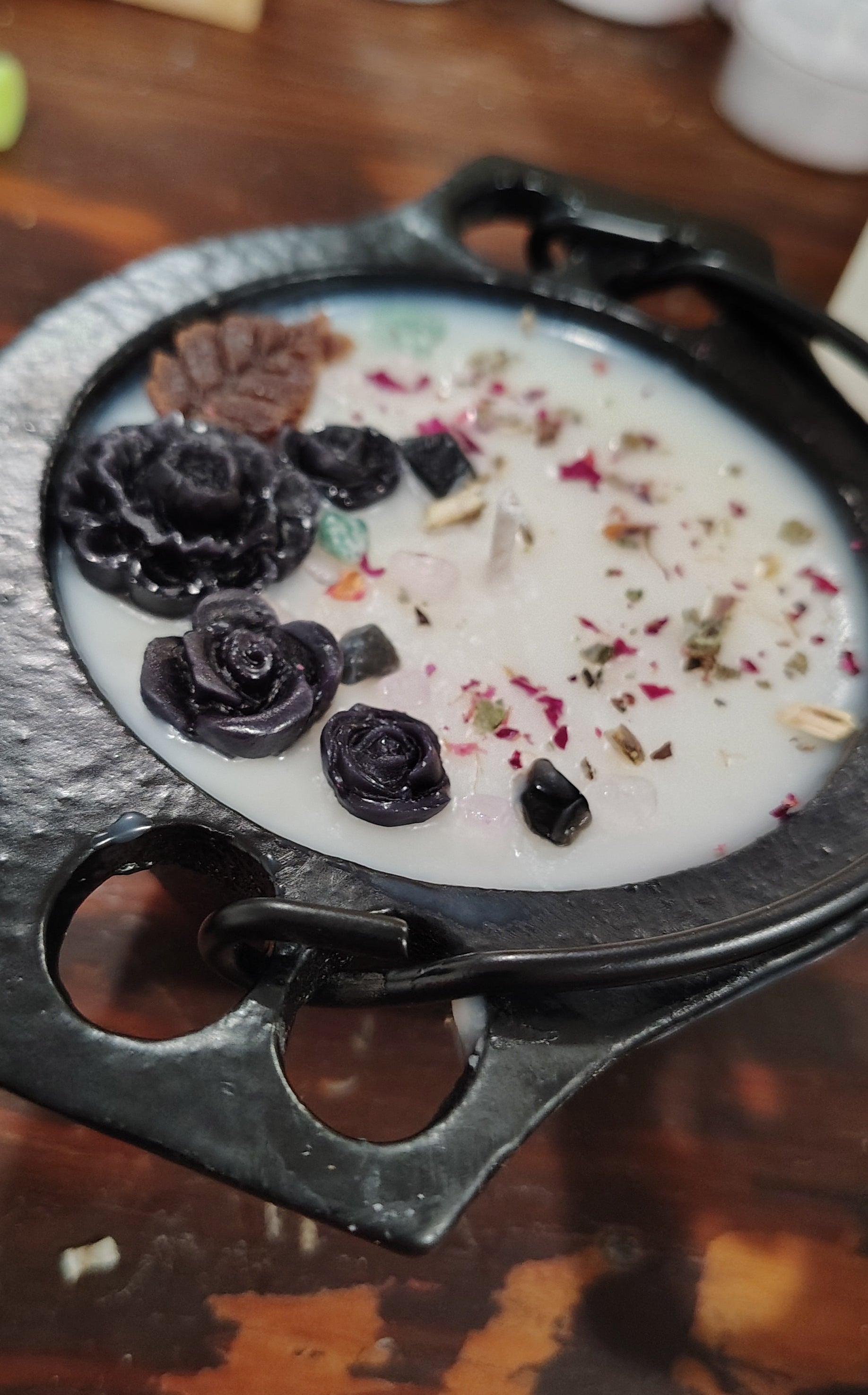 A black cast iron cauldron with decorative handles, filled with a white wax candle topped with dried botanicals and dark rose petals, set on a rustic wooden surface.