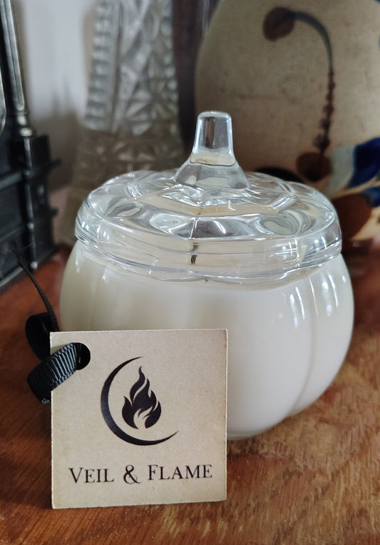 A white pumpkin-shaped candle with a clear glass lid sits on a wooden surface next to a paper tag reading 'VEIL & FLAME'. The background features blurred decorative glassware and ceramic vases.
