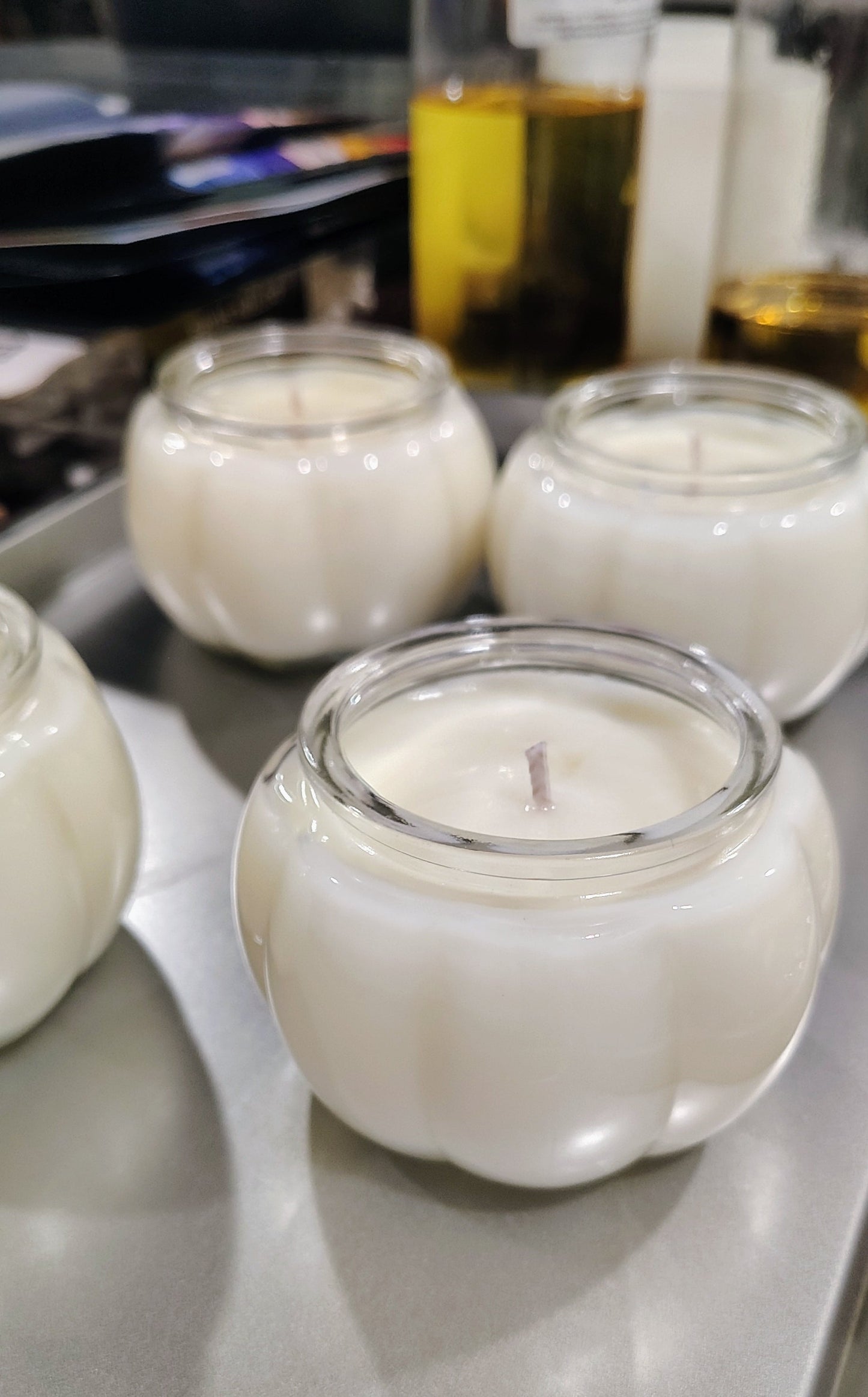 A close-up of a white wax candle in a clear glass jar with a wick, sitting on a gray metal tray with other similar candles in the background.