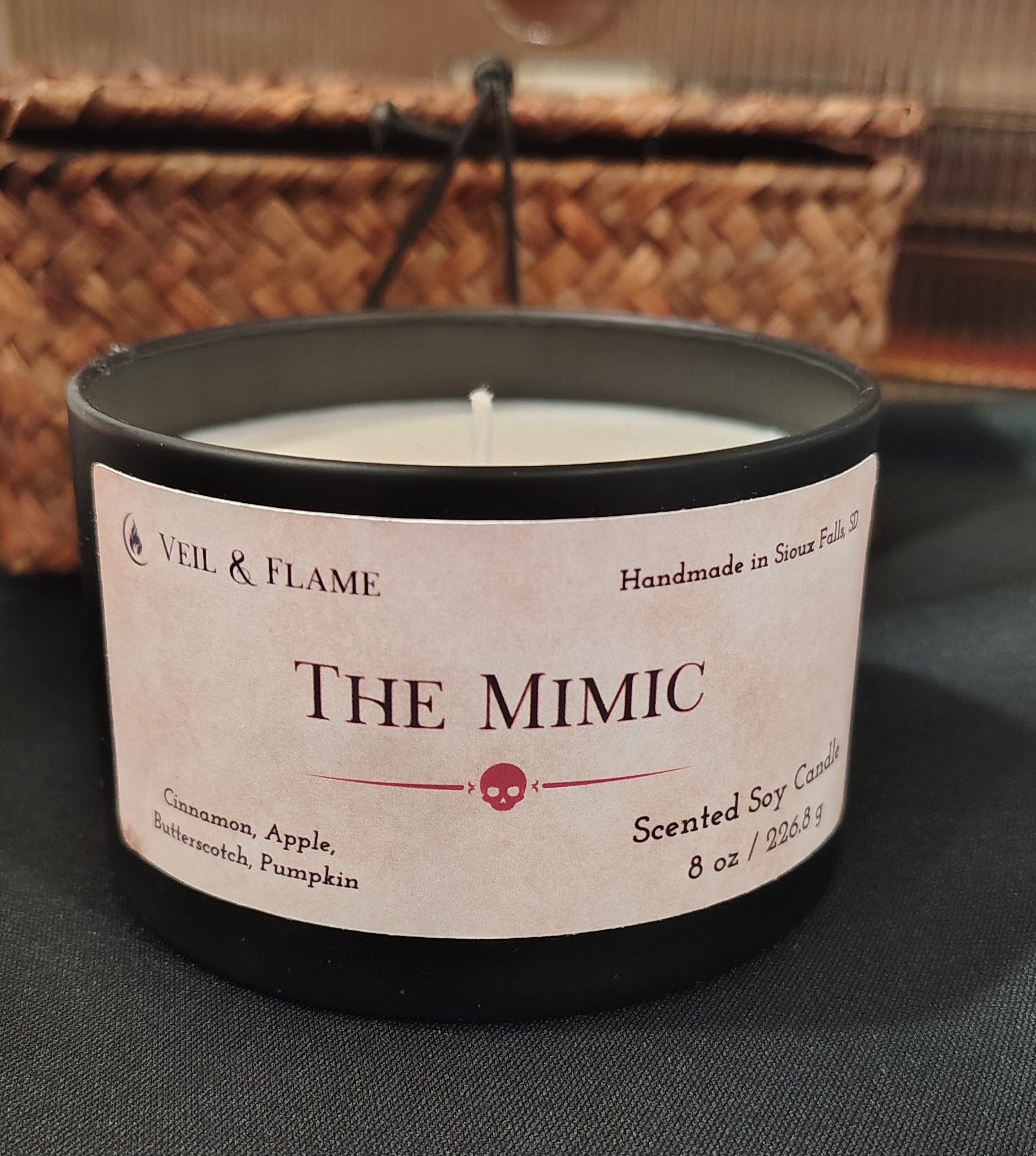 A black candle tin labeled 'THE Mimic' by Vell & Flame, placed on a dark fabric surface with a woven basket and wooden background.