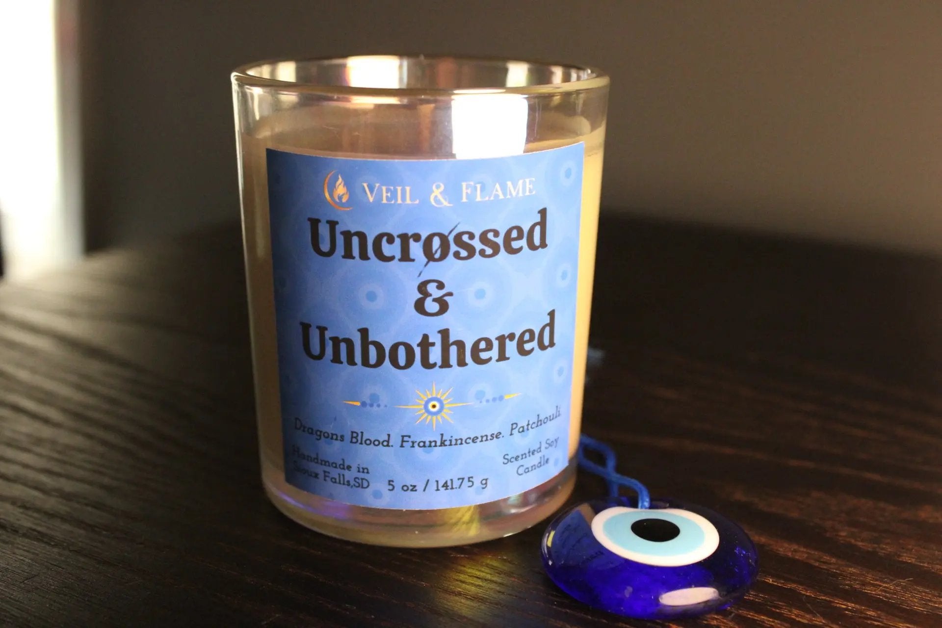 A glass jar candle labeled 'Uncrossed & Unbidden' by Veil & Flame sits on a dark wooden surface, with a blue evil eye ornament placed in front of it.
