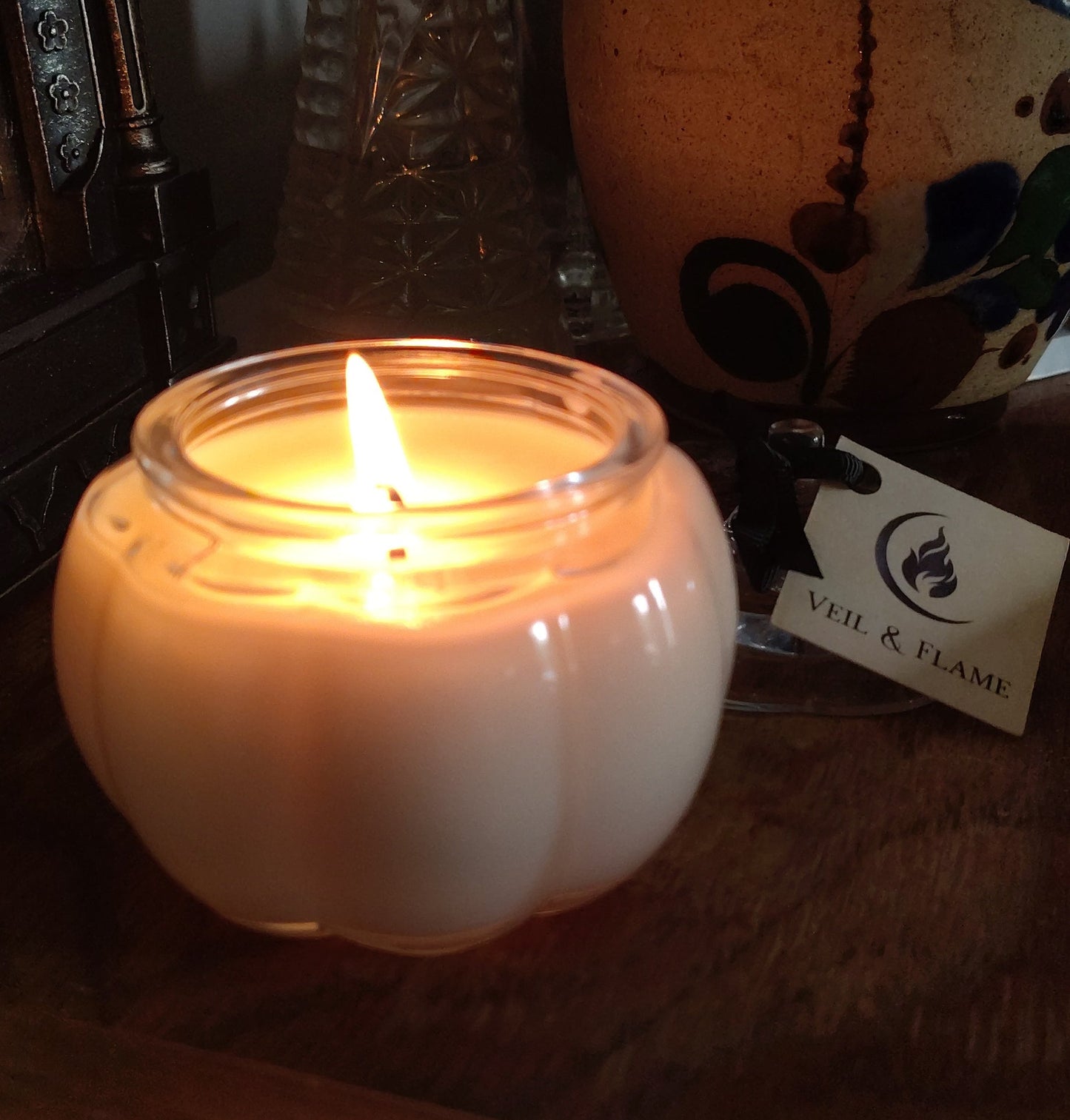 A lit white candle in a clear glass jar with a rounded pumpkin-like shape, placed on a wooden surface next to a decorative ceramic vase and a small tag.