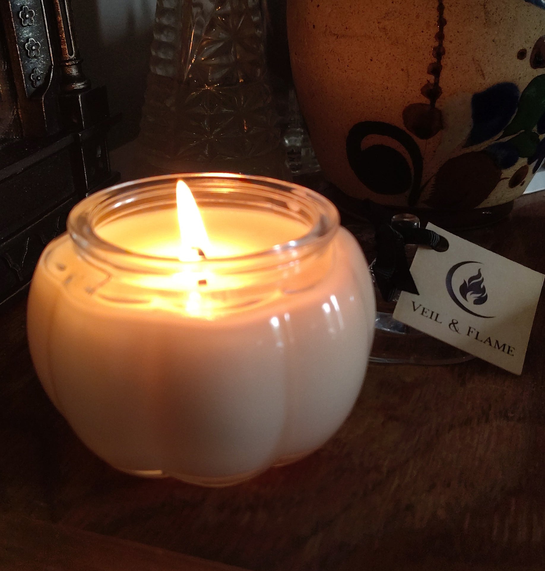 A lit white candle in a clear glass jar with a rounded pumpkin-like shape, placed on a wooden surface next to a decorative ceramic vase and a small tag.
