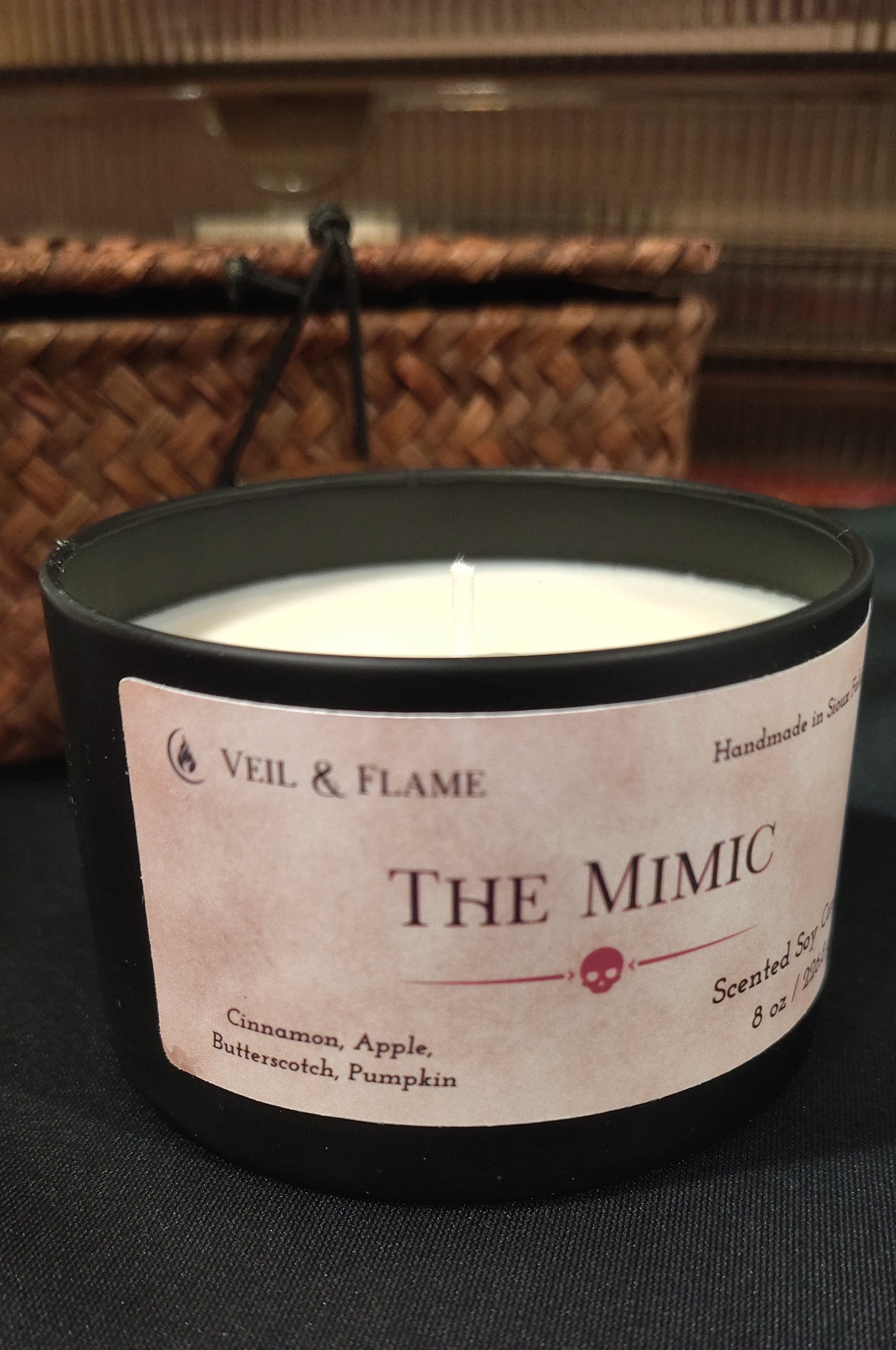 A black candle jar with a cream-colored label reading 'VEIL & FLAME THE Mimic' on a dark surface, with a woven basket blurred in the background.