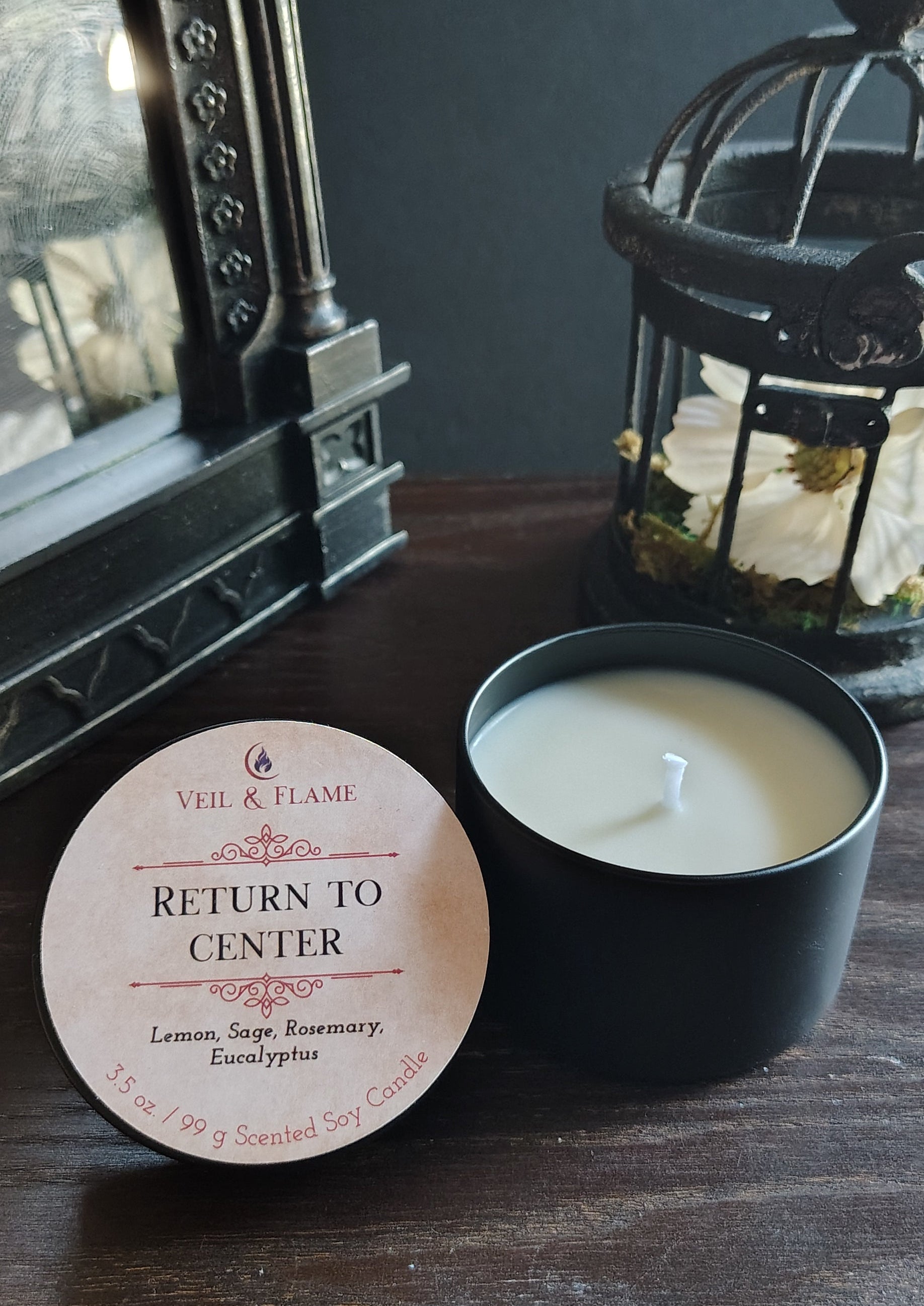 A black candle tin with a white wax candle sits on a dark wooden table next to its round cream-colored lid, which reads 'RETURN TO CENTER' and shows the brand 'Veil & FLAME'. The background features a dark gray wall, a vintage-style ornate mirror, and a black lantern.