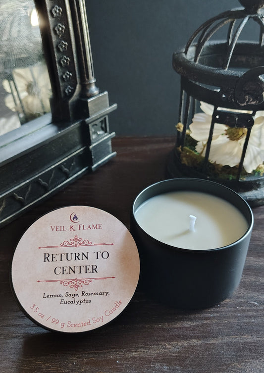 A black candle tin with a white wax candle sits on a dark wooden table next to its round cream-colored lid, which reads 'RETURN TO CENTER' and shows the brand 'Veil & FLAME'. The background features a dark gray wall, a vintage-style ornate mirror, and a black lantern.