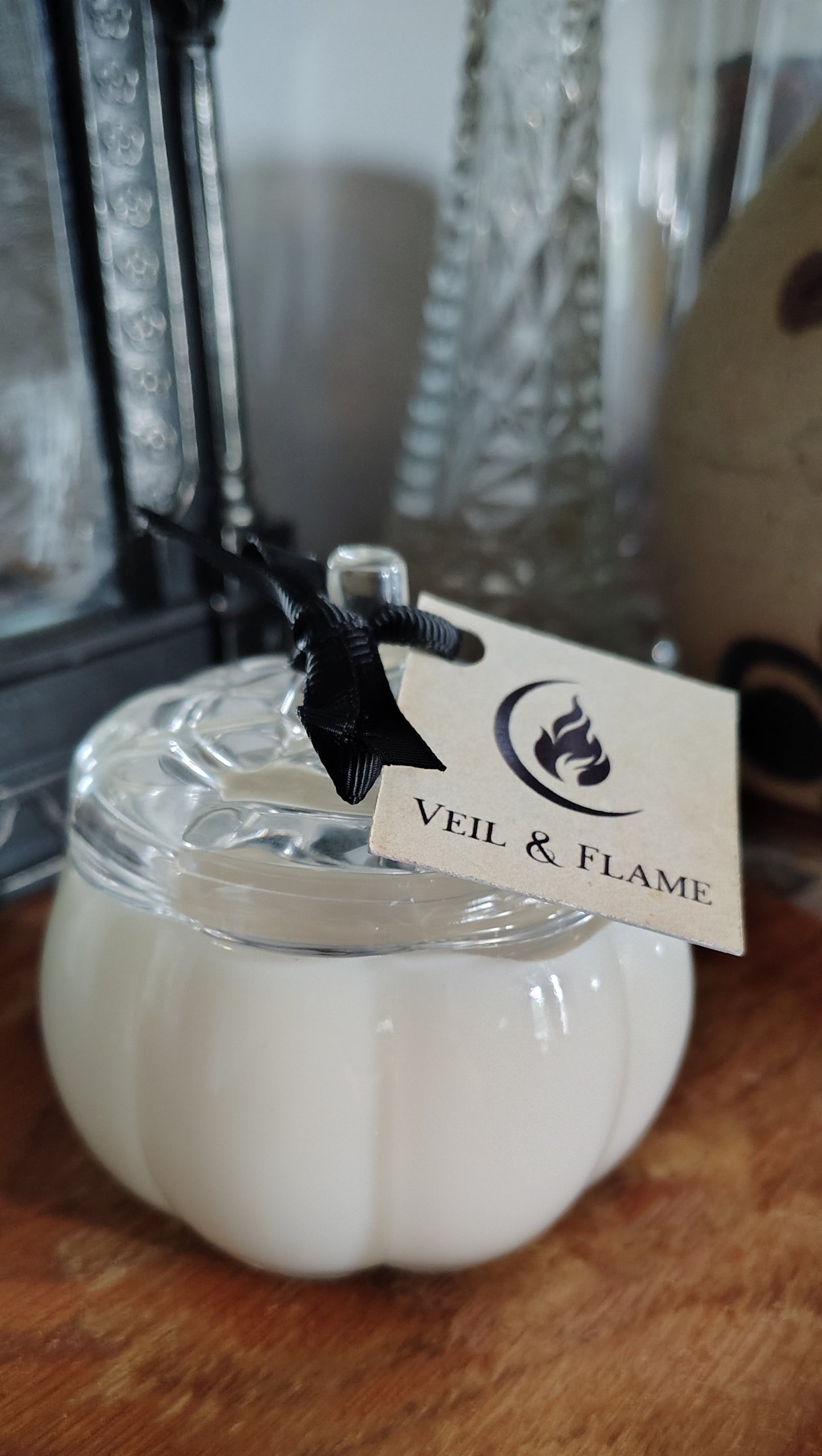 A white pumpkin-shaped candle in a clear glass jar with a glass lid, resting on a wooden surface. A black ribbon is tied to the jar, holding a paper tag that reads 'VEIL & FLAME'.