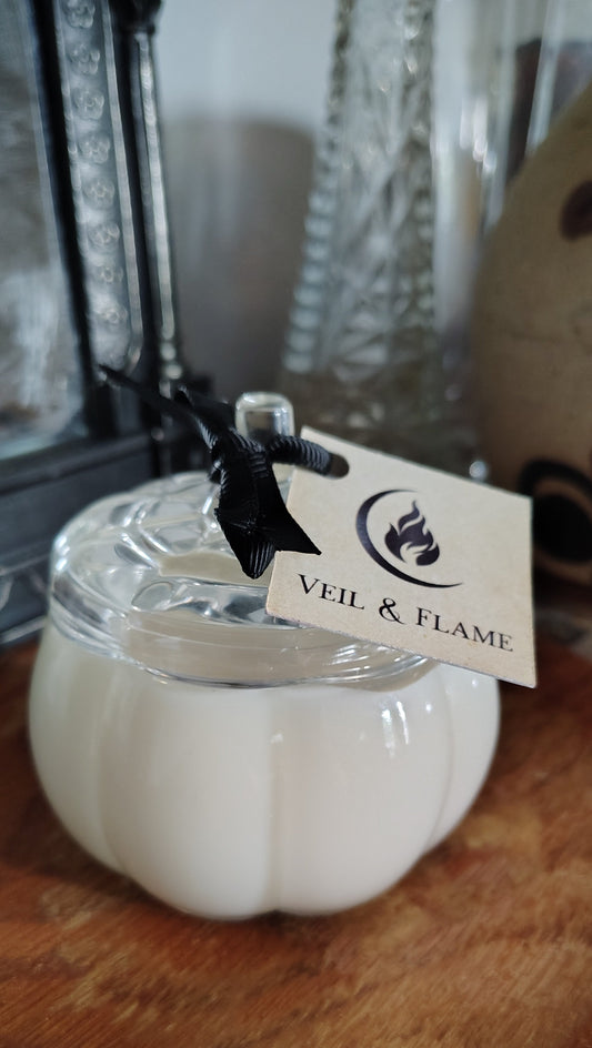 A white pumpkin-shaped candle in a clear glass jar with a glass lid, resting on a wooden surface. A black ribbon is tied to the jar, holding a paper tag that reads 'VEIL & FLAME'.