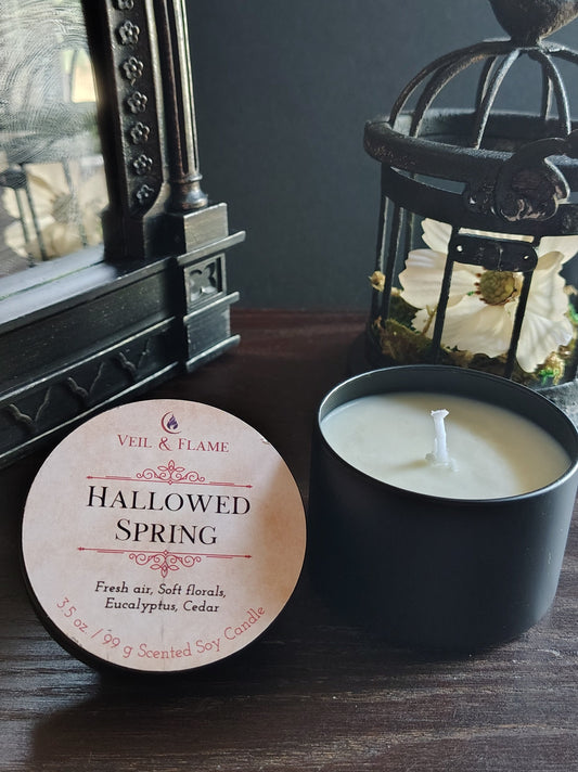 A black candle jar with cream wax sits on a dark wooden table next to its round cream-colored label, which reads 'HALLOWED SPRING' by Veil & Flame. The background features a gothic-style mirror and a birdcage lantern against a dark gray wall.