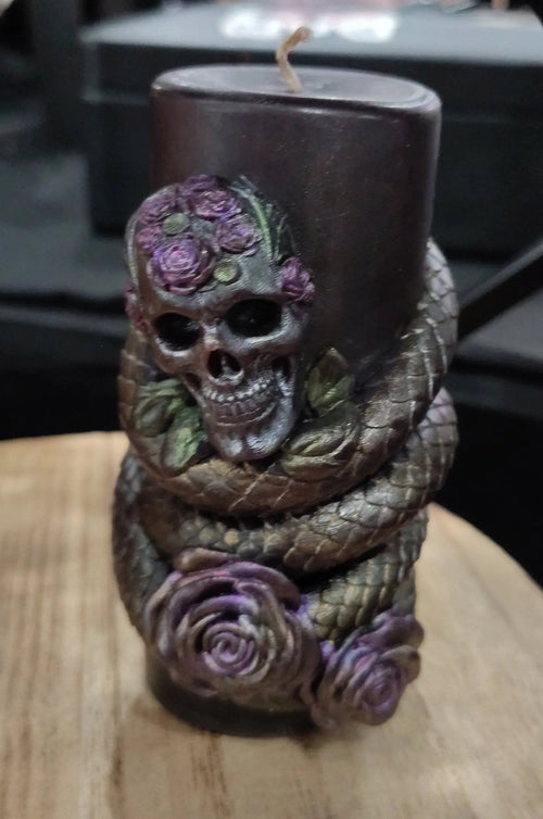 A black candle wrapped with a sculpted snake and decorated with purple roses and a skull on the front, displayed on a wooden surface.