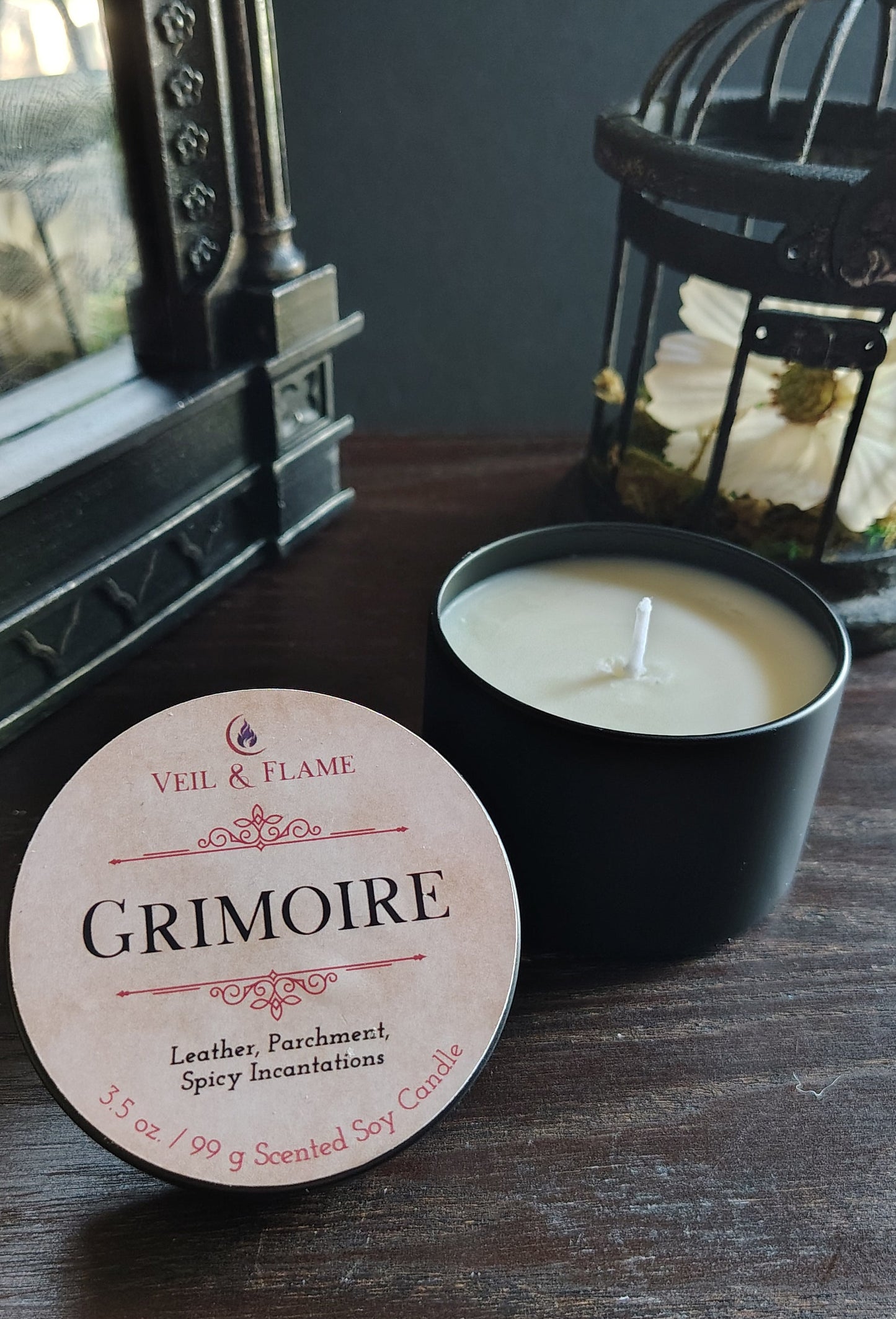 A black candle tin with a white wick sits on a dark wooden table next to its round cream-colored label, which reads 'VEIL & FLAME GRIMOIRE'. In the background, a black birdcage lantern and a dark wall are visible.