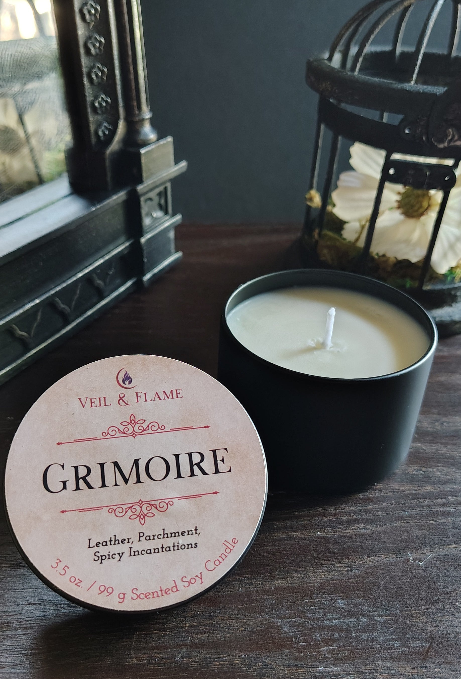 A black candle tin with a white wick sits on a dark wooden table next to its round cream-colored label, which reads 'VEIL & FLAME GRIMOIRE'. In the background, a black birdcage lantern and a dark wall are visible.