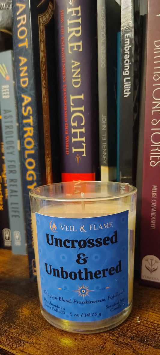 A small cylindrical candle jar with a blue label reading 'Uncrossed & Unbordered' by Veel & Flams, shown leaning against a stack of astrology and spirituality books on a wooden surface.