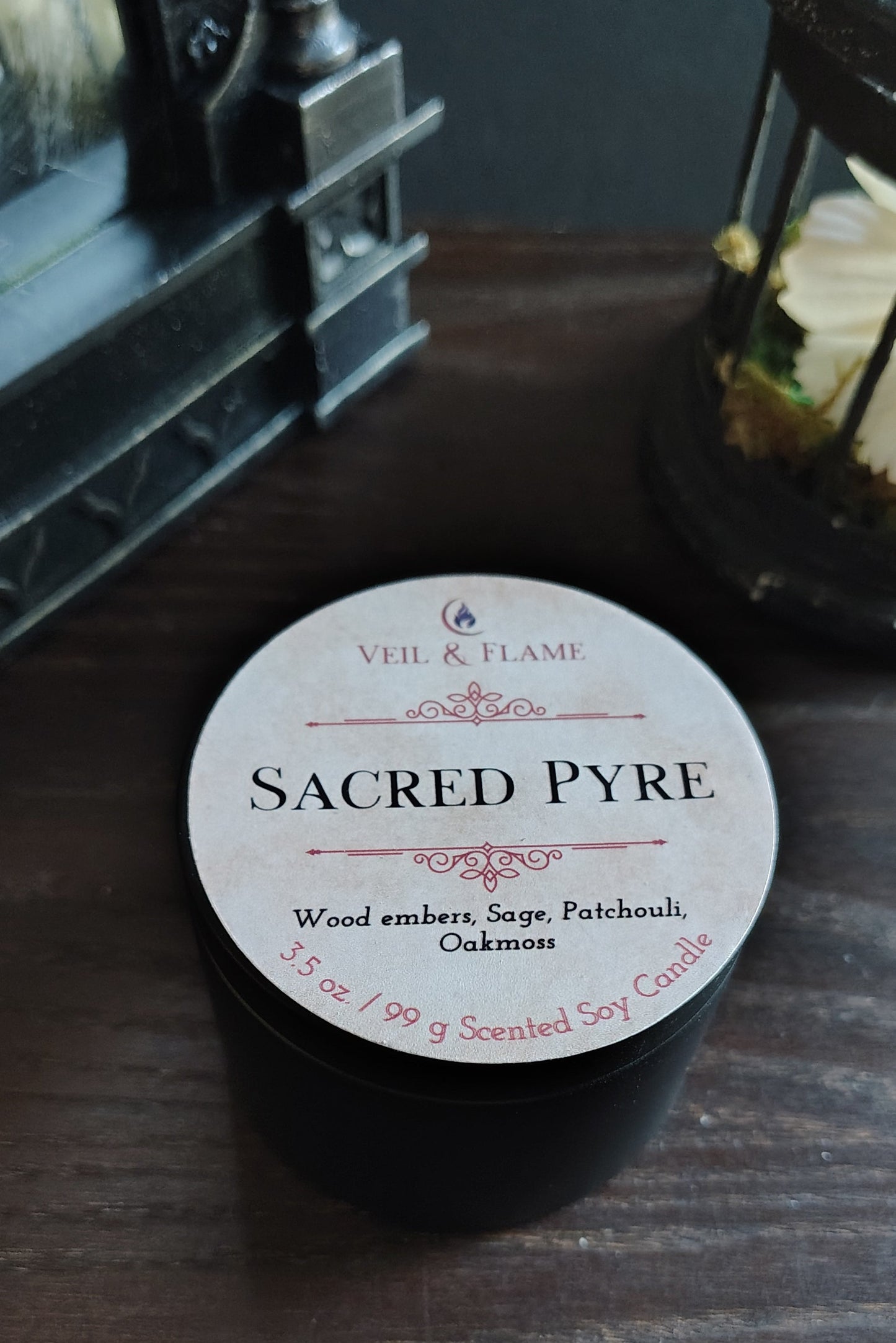 A small black jar labeled 'Sacred Pyre' by Veil & Flame, placed on a dark wooden surface with decorative lanterns in the background.