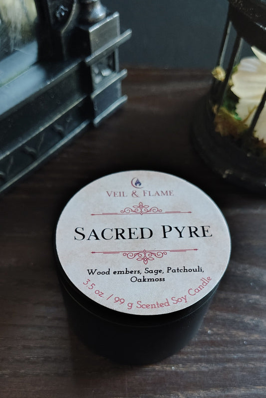 A small black jar labeled 'Sacred Pyre' by Veil & Flame, placed on a dark wooden surface with decorative lanterns in the background.