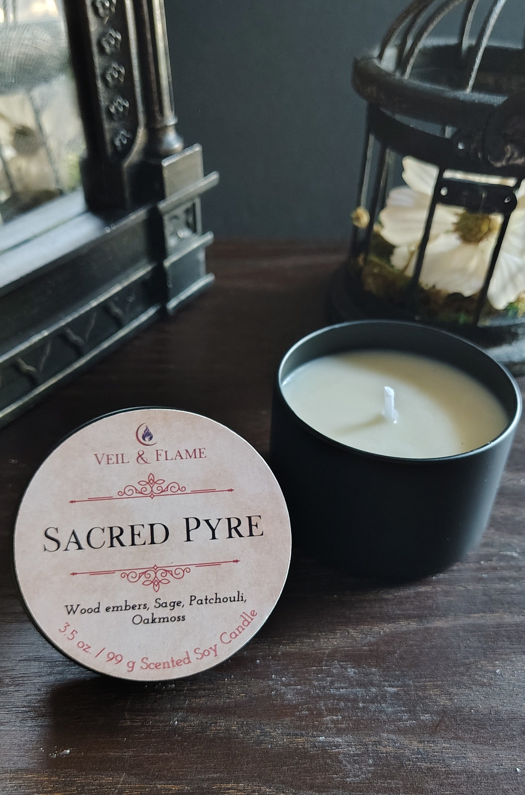 A candle product shown on a dark wooden table, with the round white paper label reading 'SACRED PYRE' in front of an open black candle jar containing white wax. Decorative lanterns and a mirror are visible in the background.