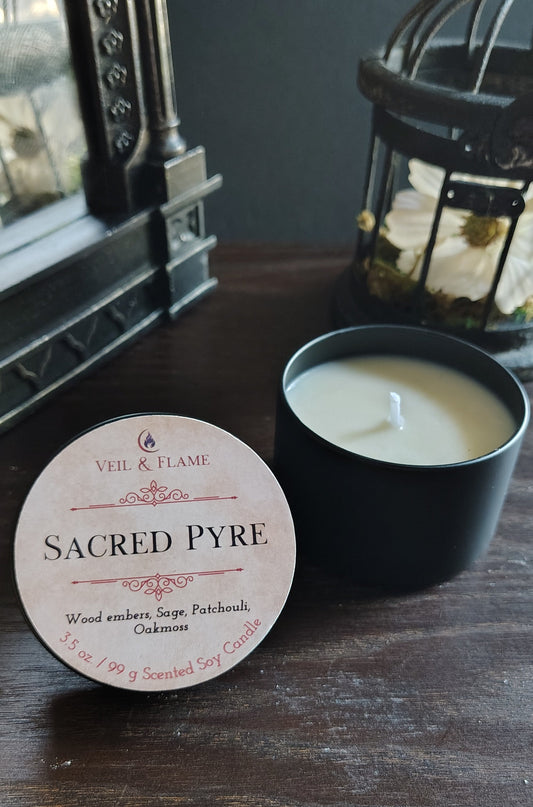 A candle product shown on a dark wooden table, with the round white paper label reading 'SACRED PYRE' in front of an open black candle jar containing white wax. Decorative lanterns and a mirror are visible in the background.